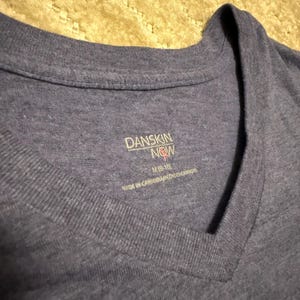 May include: Close-up of a blue Danskin Now V-neck t-shirt. The shirt has a tag with the brand name, size M (8-10), and the text "MADE IN CAMBODIA/HECHO EN CAMBOYA". The fabric appears to be a soft, heathered material.