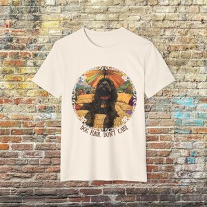 May include: A cream-coloured t-shirt featuring a graphic of a black dog lying on a bed with a colourful umbrella in the background. The text "Dog Hair, Don't Care" is printed below the graphic.