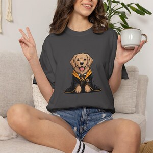 May include: Dark gray t-shirt featuring a cartoon golden retriever wearing a black and gold robe. The dog has a happy expression. The shirt is made of a soft material.