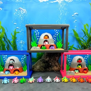 May include: A colourful underwater display with three small aquariums, each housing axolotl figurines. The aquariums have blue, grey, and pink frames. The axolotls are in various colours, including blue, pink, green, and orange, creating a lively scene.