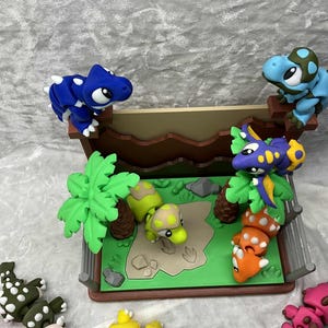 May include: A colorful dinosaur diorama featuring several small, sculpted dinosaurs in various colors, including blue, green, purple, and orange. The scene includes a miniature landscape with trees and a fence, creating a playful, imaginative setting.