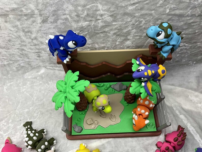 May include: A colorful dinosaur diorama featuring several small, sculpted dinosaurs in various colors, including blue, green, purple, and orange. The scene includes a miniature landscape with trees and a fence, creating a playful, imaginative setting.