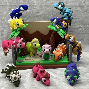 May include: A colorful diorama featuring various dinosaur figurines. The dinosaurs are in shades of blue, yellow, pink, orange, green, and purple. Some are perched on faux trees, while others stand on a green base with a brown backdrop.