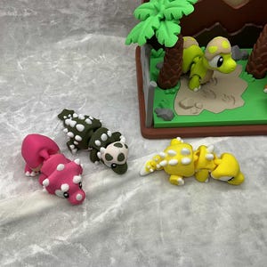 May include: A collection of colorful dinosaur figurines, including pink, green, and yellow Ankylosaurus models, and a diorama with green and orange dinosaurs. The scene is set on a light gray surface.