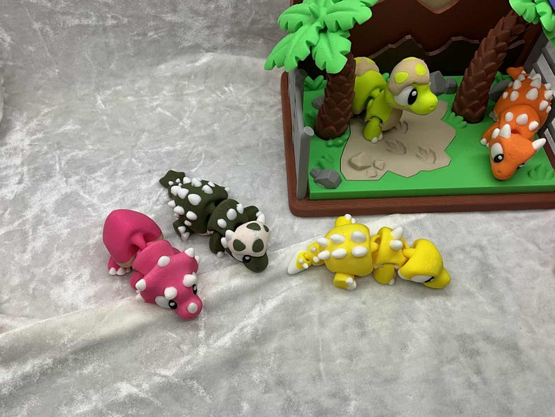 May include: A collection of colorful dinosaur figurines, including pink, green, and yellow Ankylosaurus models, and a diorama with green and orange dinosaurs. The scene is set on a light gray surface.