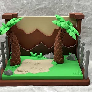 May include: A miniature diorama featuring a prehistoric scene. Two brown palm trees with green fronds stand on a green base, with a tan sand area and dinosaur footprints. A brown backdrop depicts mountains.