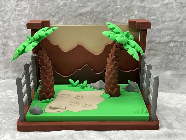 May include: A miniature diorama featuring a prehistoric scene. Two brown palm trees with green fronds stand on a green base, with a tan sand area and dinosaur footprints. A brown backdrop depicts mountains.