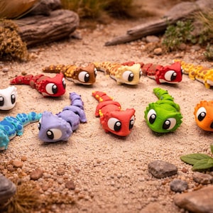 May include: A selection of brightly coloured, toy-like creatures with large eyes and segmented bodies. The creatures are in various colours, including white, blue, red, brown, yellow, green, and orange. They are arranged on a sandy surface with small rocks and foliage.