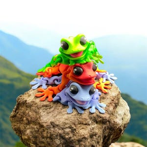 May include: A stack of three brightly coloured frog ornaments on a rock. The top frog is green, the middle one is red, and the bottom one is purple. Each frog has large black eyes and is smiling. The background shows a blurred mountain range.