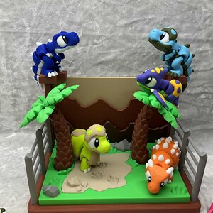 May include: A colorful diorama featuring several dinosaur figurines. The scene includes a green base with palm trees, a fence, and a sand-colored area. Dinosaurs are blue, green, orange, and purple.