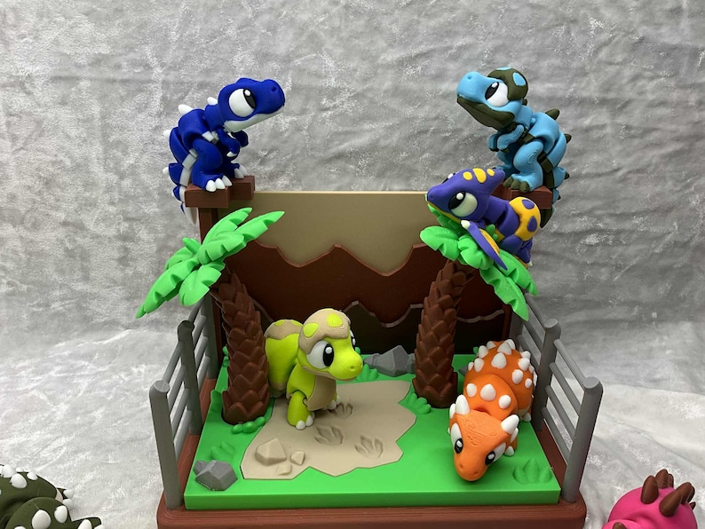 May include: A colorful diorama featuring several dinosaur figurines. The scene includes a green base with palm trees, a fence, and a sand-colored area. Dinosaurs are blue, green, orange, and purple.