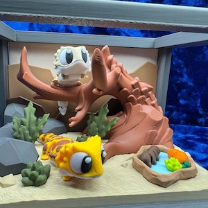 Peut inclure: Une scène de terrarium miniature avec deux lézards de dessin animé, l'un jaune et orange, l'autre blanc et beige. La scène comprend un arbre brun, des plantes vertes et une petite piscine avec de la nourriture. Le terrarium est enfermé dans un cadre gris.