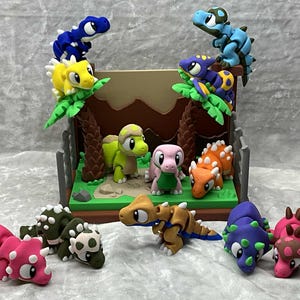 May include: A collection of colorful, handcrafted dinosaur figurines. The scene includes various dinosaurs in shades of blue, yellow, green, pink, and orange, some perched on miniature palm trees. The dinosaurs are arranged in a diorama setting with a brown backdrop.