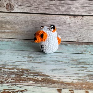 May include: A handmade, crocheted white ball with orange and black accents. The small, round object has orange ears and a black and orange nose. The toy is sitting on a weathered, light blue and brown wooden surface.