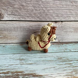 May include: A crocheted camel toy in beige with brown accents. The camel has a red halter and brown hooves. The toy is sitting on a weathered, light blue and brown wooden surface.