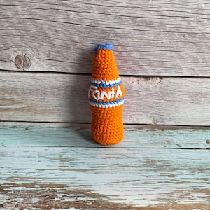 May include: Crocheted orange bottle with blue and white stripes and the word "FANTA" in white. The bottle has a blue top and is set against a wooden background. The handmade item is a unique decorative piece.
