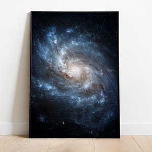 Könnte beinhalten: Ein gerahmter Druck einer Spiralgalaxie, der eine lebendige Darstellung von Blau-, Weiß- und dunklen Farbtönen zeigt. Das Kunstwerk zeigt einen Himmelskörper mit wirbelnden Armen und einem hellen zentralen Kern.