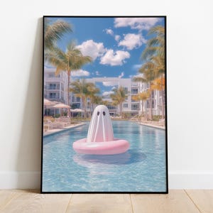 Könnte beinhalten: Gerahmter Druck, der einen Geist in einem weißen Laken auf einem rosa aufblasbaren Ring in einem Swimmingpool zeigt. Im Hintergrund sind Palmen, weiße Gebäude und ein blauer Himmel mit Wolken zu sehen. Sommerliche Szene.