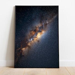 Op de afbeelding: Een ingelijste print van een hemelse scène, met een levendige weergave van sterren en kosmisch stof. Het kunstwerk heeft een donkerblauwe achtergrond met een centrale band van gouden en oranje tinten, wat een dramatisch contrast creëert. De print staat op een houten vloer.