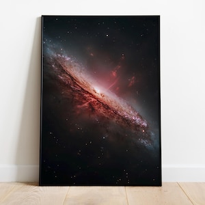 Puede incluir: Una impresión enmarcada que presenta una galaxia. La obra de arte muestra una galaxia espiral con un núcleo rojo y blanco brillante, rodeada de remolinos azules y púrpuras, sobre un fondo oscuro y lleno de estrellas. La impresión está en un marco negro.
