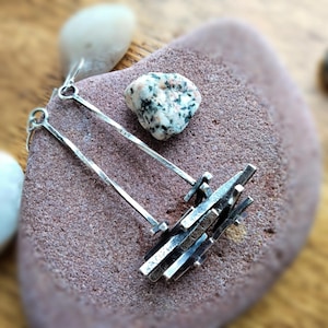May include: A pair of silver earrings with a modern, geometric design. Each earring features a long, slender bar with a textured finish, connected to a cluster of smaller, rectangular shapes. The earrings are displayed on a reddish-brown stone, with a small rock in the background.