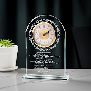 May include: A clear glass desk clock with a gold-colored clock face and Roman numerals. The clock has a decorative wreath design and the text "NEVER UNDERESTIMATE The Difference YOU MADE AND THE LIVES You Touched Happy Retirement!"