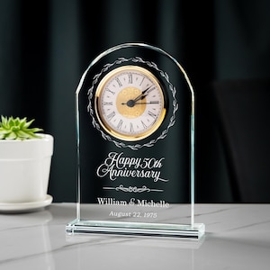 May include: A clear glass clock with a gold-colored clock face and Roman numerals. The clock is engraved with the words "Happy 50th Anniversary," the names "William & Michelle," and the date "August 22, 1975."