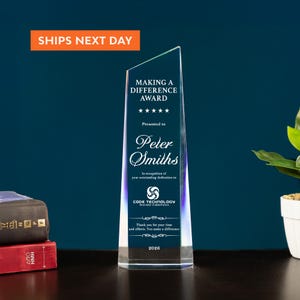 May include: A clear, wedge-shaped glass award featuring an angled top. The front is engraved with the text Making a Difference Award, five stars, Peter Smiths, Code Technology Innovator in Electronics, and the year 2026 in white lettering.