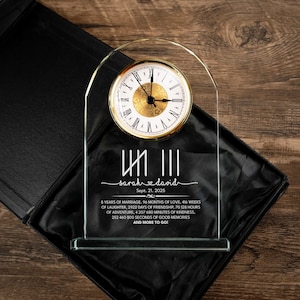 May include: A clear glass clock with a gold-colored clock face and Roman numerals. The clock is engraved with the names "Sarah & David" and the date "Sept. 21, 2025". The base includes text about years of marriage, months of love, and more.