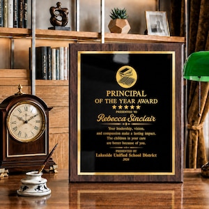 May include: A rectangular award plaque with a dark wood frame and a black center panel featuring gold text that reads Principal of the Year Award, presented to Rebecca Sinclair by Lakeside Unified School District in 2026, with a book icon.