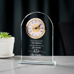 May include: A clear glass desk clock with a gold-tone clock face and Roman numerals. The clock has an arched top and a rectangular base. Text on the clock reads "Never Underestimate The Difference You Made and The Lives You Touched" and includes a name and law firm.