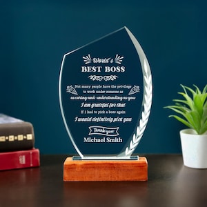 May include: A clear glass award with the text "World's Best Boss" and a personalized message. The award is mounted on a rectangular wooden base. A decorative leaf design is on the right side of the award.