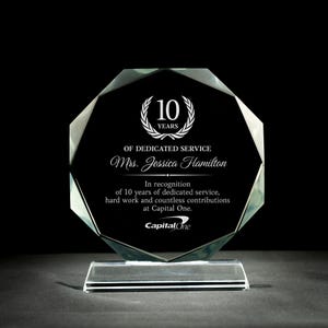 May include: A clear glass octagonal award plaque with a black background and white text. The plaque features a laurel wreath with the number 10 in the center and the text "Years" below. The text "Of Dedicated Service" is below the wreath, followed by the name "Mrs. Jessica Hamilton". The text "In recognition of 10 years of dedicated service, hard work and countless contributions at Capital One." is below the name. The Capital One logo is at the bottom of the plaque.