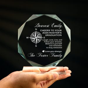 May include: A clear glass award with a compass design and the text "Dearest Emily, CHEERS TO YOUR 2025 MASTER'S GRADUATION Laugh, Love, Live, and Follow your dreams. Believe in yourself and remember to stay awesome! Love you always. The Foster Family"