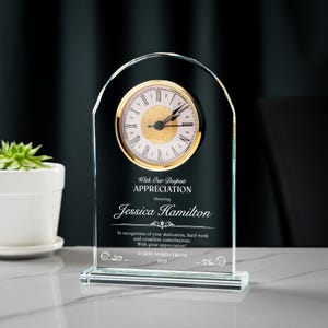 May include: A clear glass clock with a gold-colored clock face and Roman numerals. The clock has a black background with text that reads "With Our Deepest Appreciation" and "Jessica Hamilton". The clock is approximately 6 inches tall.