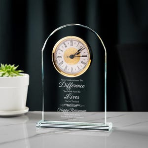 May include: A clear glass clock with a gold-colored clock face and Roman numerals. The clock has an arched top and a rectangular base. Text on the clock reads "Never Underestimate The Difference You Made And The Lives You've Touched. Happy Retirement."