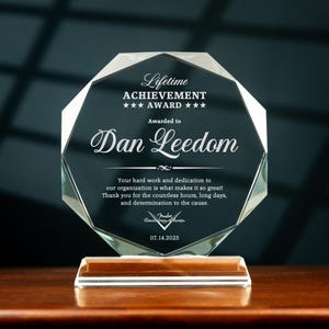 May include: A clear, faceted glass award with the text "Lifetime ACHIEVEMENT AWARD" and "Awarded to Dan Leedom." The base is rectangular and clear. The text includes a date and a signature.