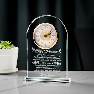 May include: A clear glass desk clock with a gold-colored clock face and Roman numerals. The clock has the words "Happy Retirement!" and a personalized message engraved on the glass. The clock is approximately 6 inches tall.