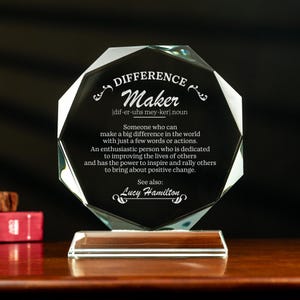 May include: A clear glass award with a beveled octagonal shape. The award features the word "Difference Maker" with a definition and the name "Lucy Hamilton".