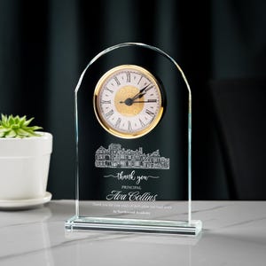May include: A clear glass desk clock with a gold-colored clock face and Roman numerals. The clock has a decorative etched design of a building and the words "thank you PRINCIPAL Ava Collins" and other text.