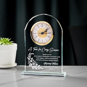 May include: Clear glass clock with a gold-tone clock face and Roman numerals. The clock has an arched top and a rectangular base. Text on the clock reads "A Time for Every Season" and includes a personalized message and the year 2025.