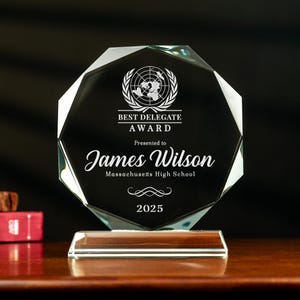 May include: A clear, faceted glass award with a wooden base. The award features the text "BEST DELEGATE AWARD" and "James Wilson" with the year "2025". The award is set against a dark background.