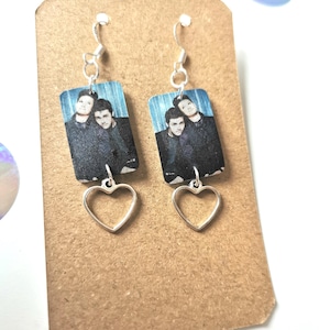 May include: A pair of dangle earrings with a silver heart charm and a rectangular photo charm featuring two people. The photo charm has a blue background and the people are wearing black clothing.