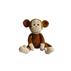 Puede incluir: Un juguete de mono de crochet marrón con manos y pies de crochet blanco. El mono tiene ojos negros y una sonrisa cosida.