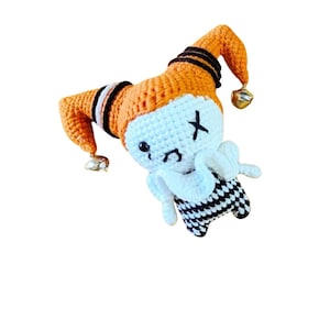 Peut inclure: Un pantin de jester crocheté avec un visage blanc, des yeux noirs et un X noir sur un œil. Le pantin porte une tenue à carreaux noirs et blancs et a un chapeau orange avec des rayures noires et des cloches.