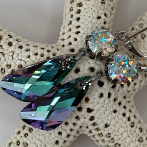 May include: A pair of earrings with iridescent, teardrop-shaped crystals and round, clear crystals. The earrings have silver-colored metal findings and are displayed on a white, textured starfish.