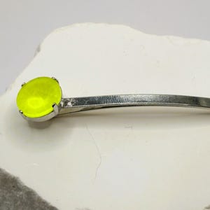 May include: A silver-toned bobby pin featuring a bright neon yellow gemstone. The bobby pin is a simple, elegant design, perfect for securing hair. The gemstone is round and set in a silver bezel.