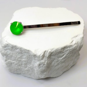 May include: A silver-toned hair barrette with a bright green, round gemstone. The barrette is set on a white, textured surface. The gemstone is a vibrant, eye-catching accent.