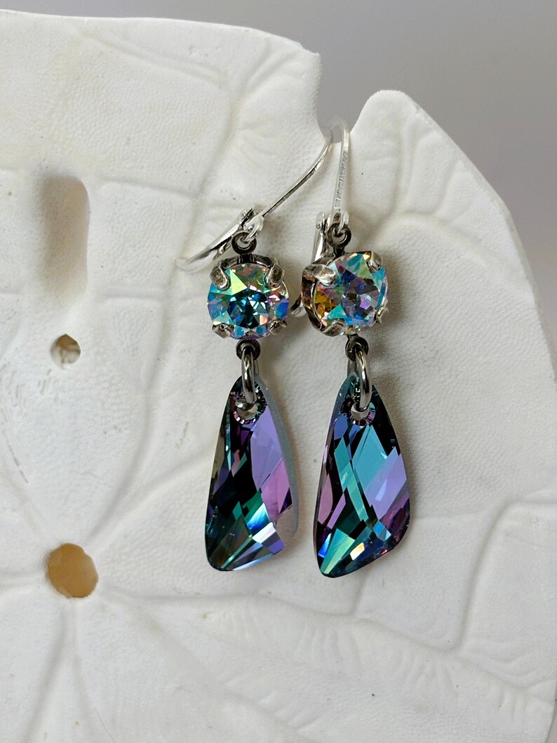 May include: A pair of iridescent crystal earrings. Each earring features a round, multi-colored crystal at the top, connected to a teardrop-shaped crystal pendant. The crystals reflect shades of blue, purple, and green, with silver-tone ear wires.