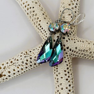 May include: A pair of silver-toned earrings with iridescent, teardrop-shaped crystals. The earrings have a lever-back closure and are displayed on a white starfish. The crystals reflect shades of teal, purple, and green.